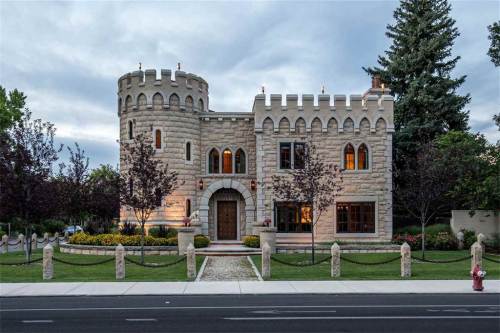 A large stone castle-style house with turrets, surrounded by trees and a well-maintained garden, under a cloudy sky.