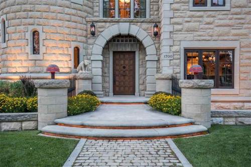 A stone house entrance with a wooden door, lanterns, and landscaped flower beds along a paved walkway.