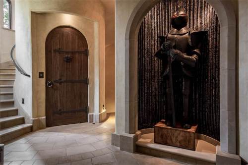 A grand entryway featuring a wooden door, a knight statue, and a staircase with stone flooring.