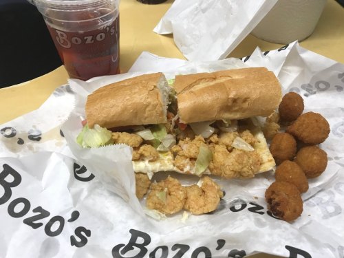 A sandwich filled with shrimp and lettuce, accompanied by hushpuppies and a drink, all on Bozo's Deli paper.