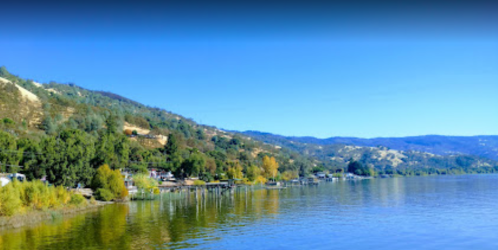 Scenic view of a calm lake surrounded by green hills and autumn-colored trees under a clear blue sky.