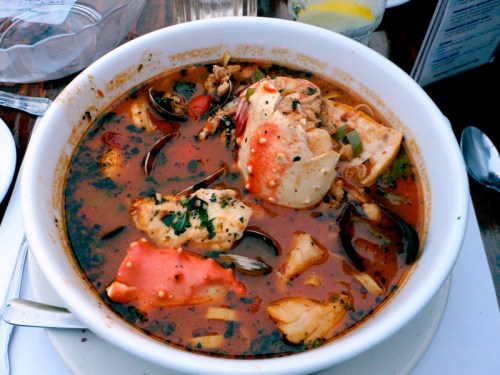 A bowl of spicy seafood soup with crab, fish, and herbs, served in a rich, red broth.
