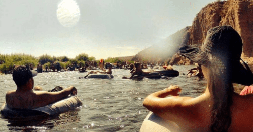 People relaxing on inner tubes in a river, surrounded by greenery and rocky cliffs under a sunny sky.