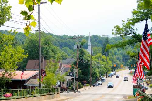 A small town street lined with trees, flags, and a church in the background, with cars driving along the road.