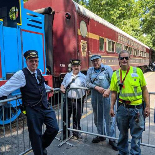 Four train conductors pose in front of a vintage train, with a blue engine and red passenger car in the background.