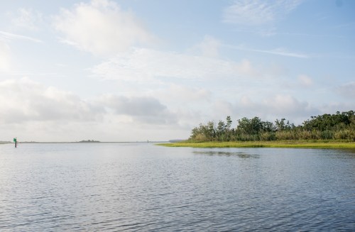 A serene waterway with gentle waves, lush greenery on the shore, and a clear blue sky with scattered clouds.