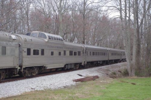 A vintage silver train car parked on a gravel track, surrounded by bare trees and grass.