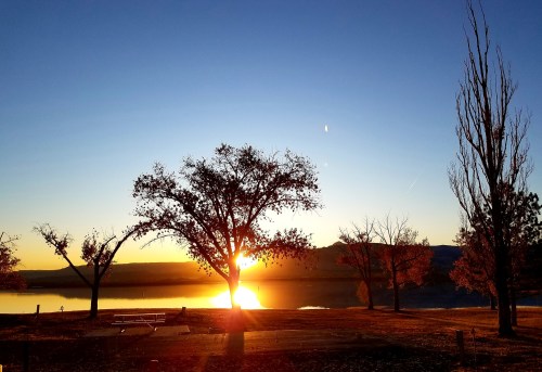 A serene sunset over a lake, with silhouetted trees and a clear sky reflecting warm colors.