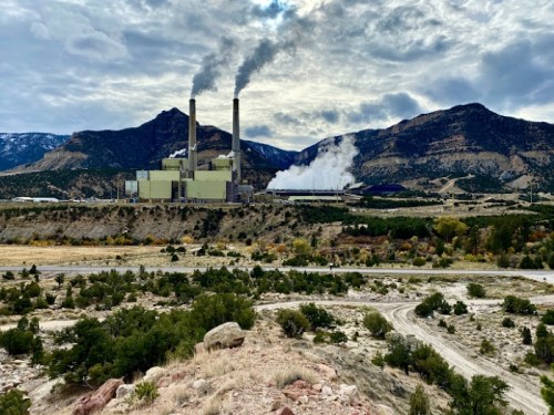 A power plant with two smokestacks emits smoke, set against a mountainous landscape under a cloudy sky.