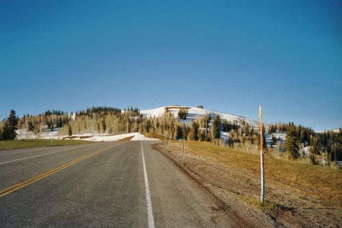 A clear road leads towards a snow-covered hill, surrounded by trees under a bright blue sky.
