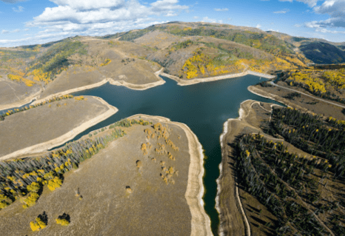 Aerial view of a winding river surrounded by autumn-colored hills and trees under a blue sky with scattered clouds.