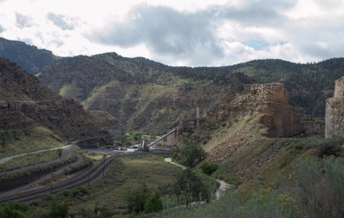 A scenic view of mountains and a valley with industrial structures and railway tracks under a cloudy sky.