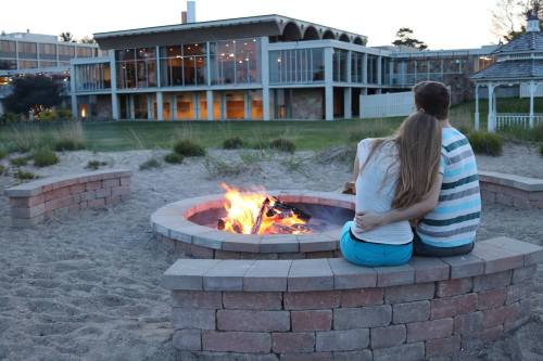 A couple sits by a fire pit on the beach, enjoying the sunset with a modern building in the background.