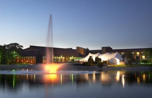 A serene evening scene of a building by a lake, featuring a fountain illuminated in warm colors and a tent-like structure.