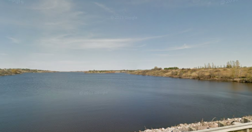 A serene view of a calm river surrounded by grassy banks and distant trees under a clear blue sky.