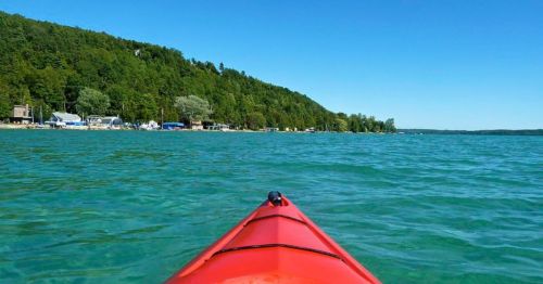 A red kayak glides through clear blue water, with a lush green shoreline in the background under a bright blue sky.