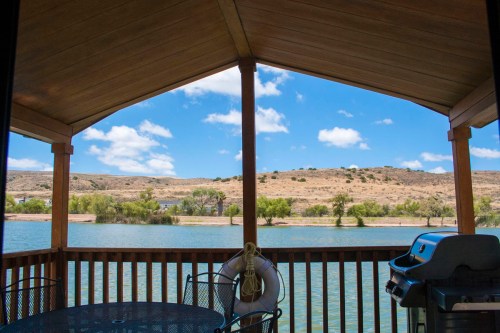View from a wooden deck overlooking a calm lake, with hills and blue sky in the background. A grill is visible.