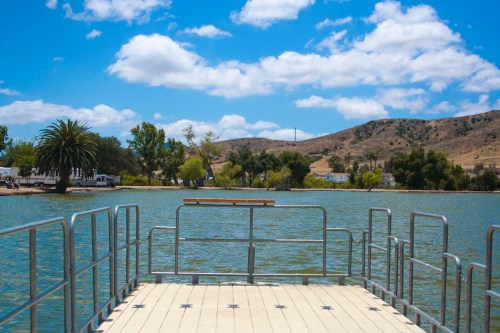 A view from a dock overlooking a calm lake, surrounded by trees and hills under a blue sky with fluffy clouds.
