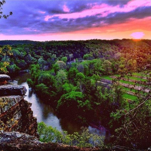 A vibrant sunset over a lush green landscape and winding river, viewed from a rocky overlook.