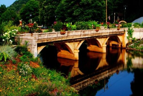 A stone bridge arches over a calm river, surrounded by lush greenery and colorful flower pots.