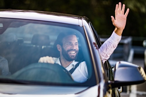 A smiling man in a car waves from the driver's seat, wearing a white shirt and looking happy.