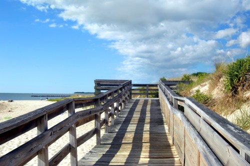 A wooden boardwalk leads to a sandy beach under a blue sky with scattered clouds.