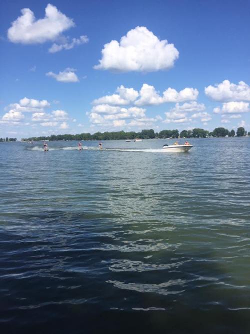 A boat speeds across a calm lake under a blue sky with fluffy clouds, surrounded by green trees in the distance.