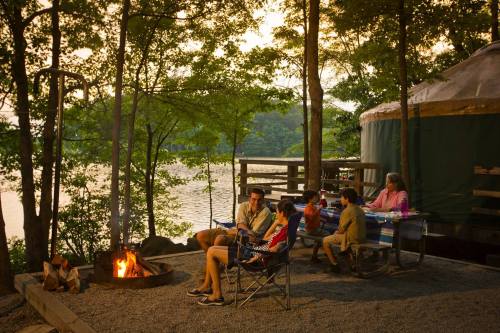 A family gathers around a campfire by a lake, enjoying a cozy evening near a yurt surrounded by trees.