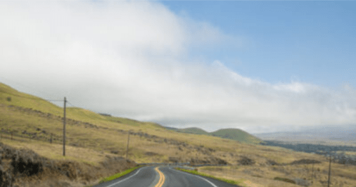 A winding road stretches through a grassy landscape under a partly cloudy sky.