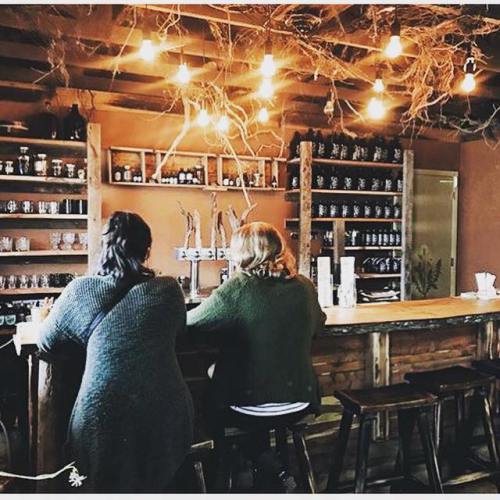 Two people sit at a rustic bar, surrounded by shelves of jars and warm lighting in a cozy setting.