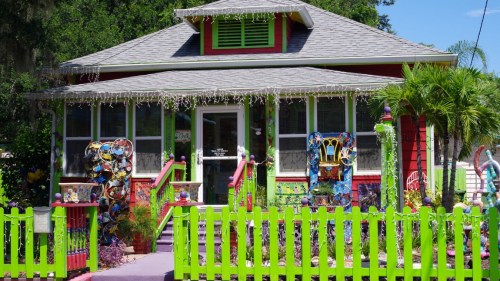 Colorful house with vibrant green fence, decorated porch, and artistic details, surrounded by lush greenery.