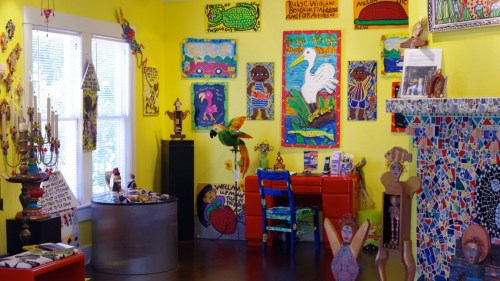 Colorful room filled with vibrant artwork, sculptures, and decorative items against bright yellow walls.