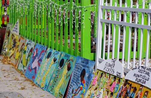 Colorful paintings displayed along a bright green fence, with festive lights in the background.