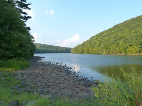 A serene lake surrounded by lush green hills under a clear blue sky, with rocky shores and gentle reflections on the water.