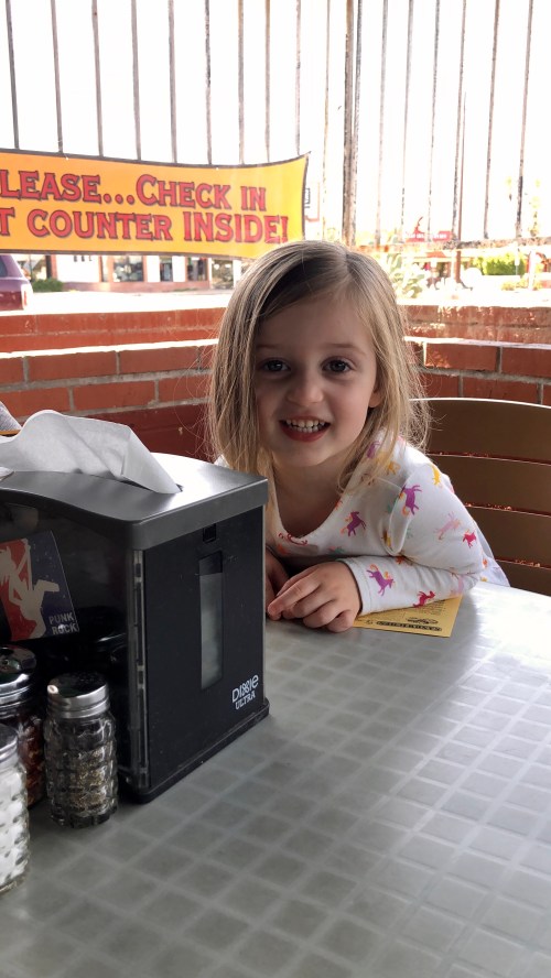 A young girl with long hair smiles at a table with a tissue box and condiments in a casual dining setting.