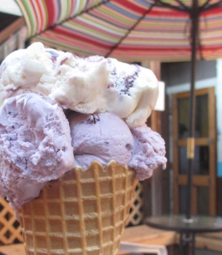 A close-up of a waffle cone filled with three scoops of ice cream: purple, white, and a mix of both, under a colorful umbrella.