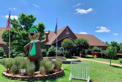 A large cartoon bear statue waves in front of a log cabin building with flags and greenery under a blue sky.