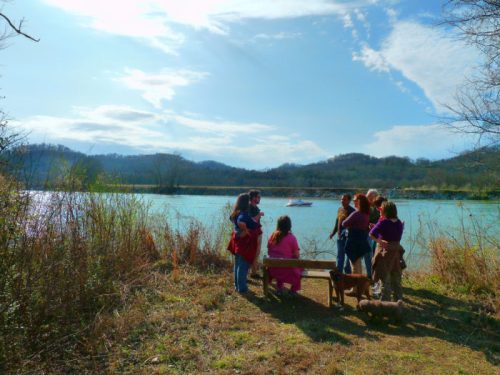 A group of people stands by a river, with mountains in the background and a boat on the water. A dog is nearby.