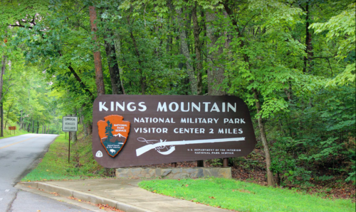 Sign for Kings Mountain National Military Park, indicating visitor center is 2 miles ahead, surrounded by greenery.