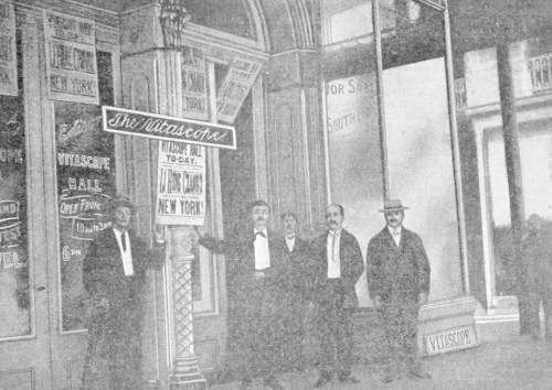 Five men in formal attire stand outside a vintage storefront with signs, including one for "New York."