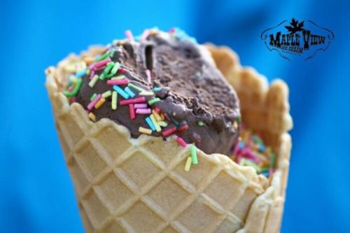 A close-up of a waffle cone filled with chocolate ice cream topped with colorful sprinkles against a blue background.