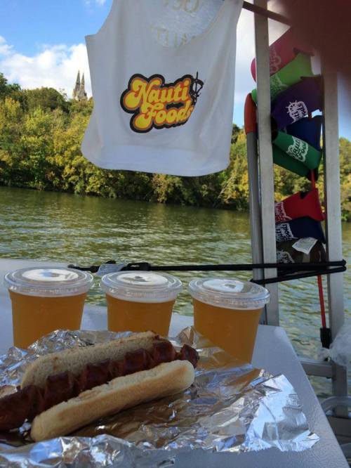 A boat scene featuring a hot dog, drinks, and a tank top with "Nauti Food" by a river surrounded by trees.