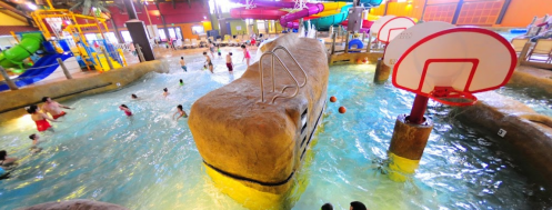 Indoor water park featuring slides, a rock formation, and children playing in the pool with basketball hoops.