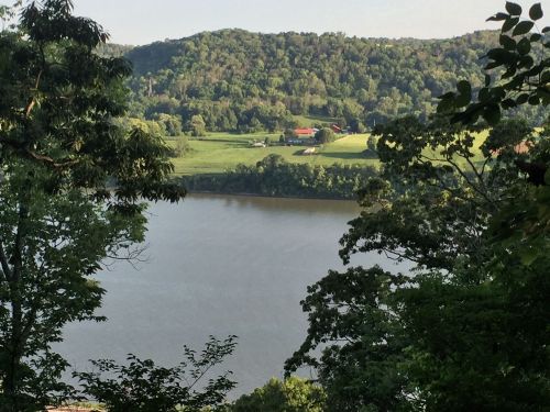 A serene river view surrounded by lush green hills and trees, with a glimpse of farmland in the distance.