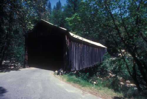 A rustic wooden covered bridge surrounded by trees, with a gravel road leading up to its entrance.