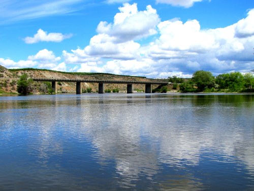 A serene river scene with a bridge spanning across, surrounded by lush greenery and fluffy clouds reflecting in the water.