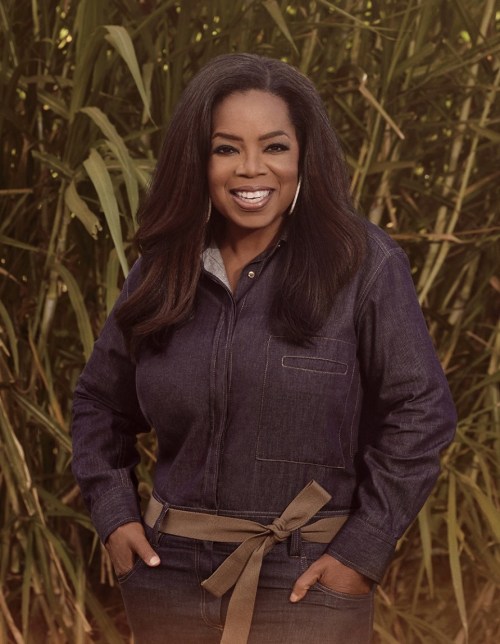 A smiling woman in a denim jacket stands outdoors, surrounded by tall green plants, exuding warmth and confidence.
