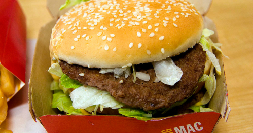 A close-up of a sesame seed bun burger with lettuce and a beef patty in a red takeout box.