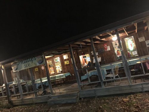A rustic wooden restaurant with a porch, dimly lit at night, featuring checkered tablecloths and a welcoming atmosphere.