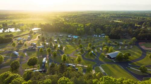 Aerial view of a lush campground with RVs, trees, and a sunset over green fields and winding paths.